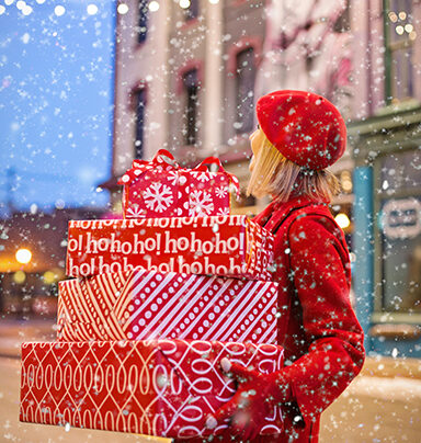 woman on sidewalk standing in falling snow carrying gift-wrapped Christmas presents